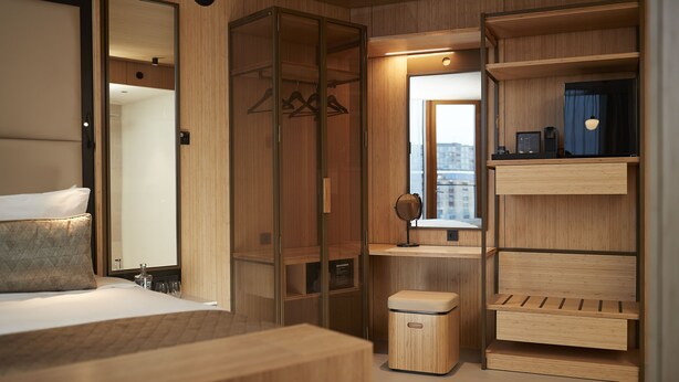 Quarto de hotel moderno com cama, guarda-roupa de madeira, um banquinho e um espelho.