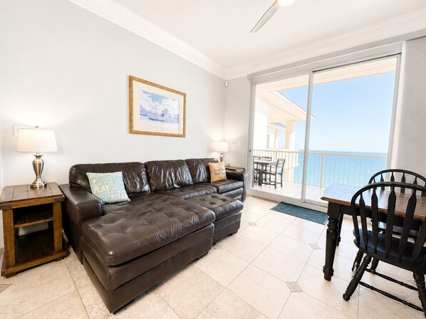A living room with a brown leather sofa, two side tables with lamps, a framed picture on the wall, and a balcony with a view of the sea.