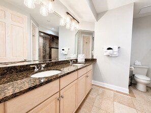 A bathroom with a granite countertop, two sinks, a large mirror, and a toilet.