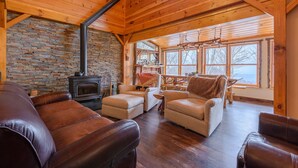 A cozy cabin interior with a stone wall, a fireplace, leather sofas, and a dining area with large windows. - Luxury Mountain Cabin with Views (Banner Elk)