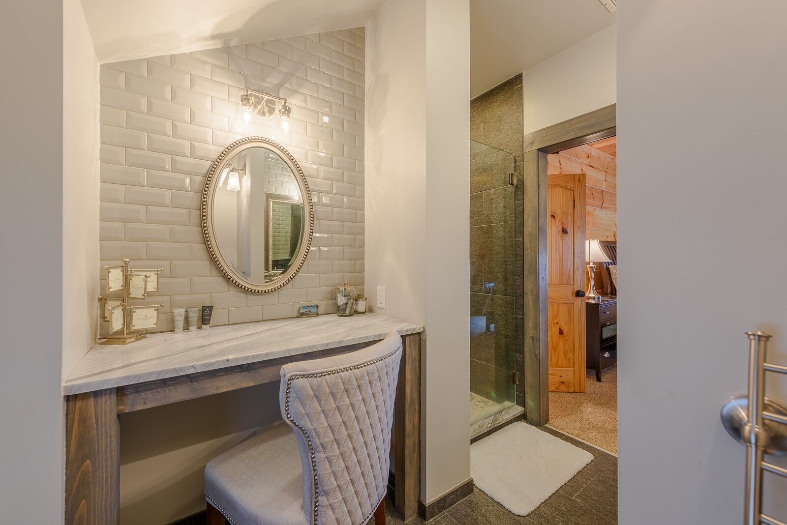 A bathroom with a white tiled backsplash, a round mirror with a gold frame, a vanity with a marble countertop, and a glass shower enclosure.