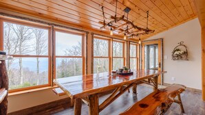 A large wooden dining table with benches, large windows, and a rustic ceiling with exposed beams and hanging light fixtures. - Luxury Mountain Cabin with Views (Banner Elk)