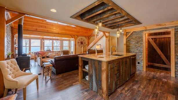 A rustic cabin interior with a central island, wooden floors, a fireplace, and a large window overlooking a forested area. - Luxury Mountain Cabin with Views (Banner Elk)