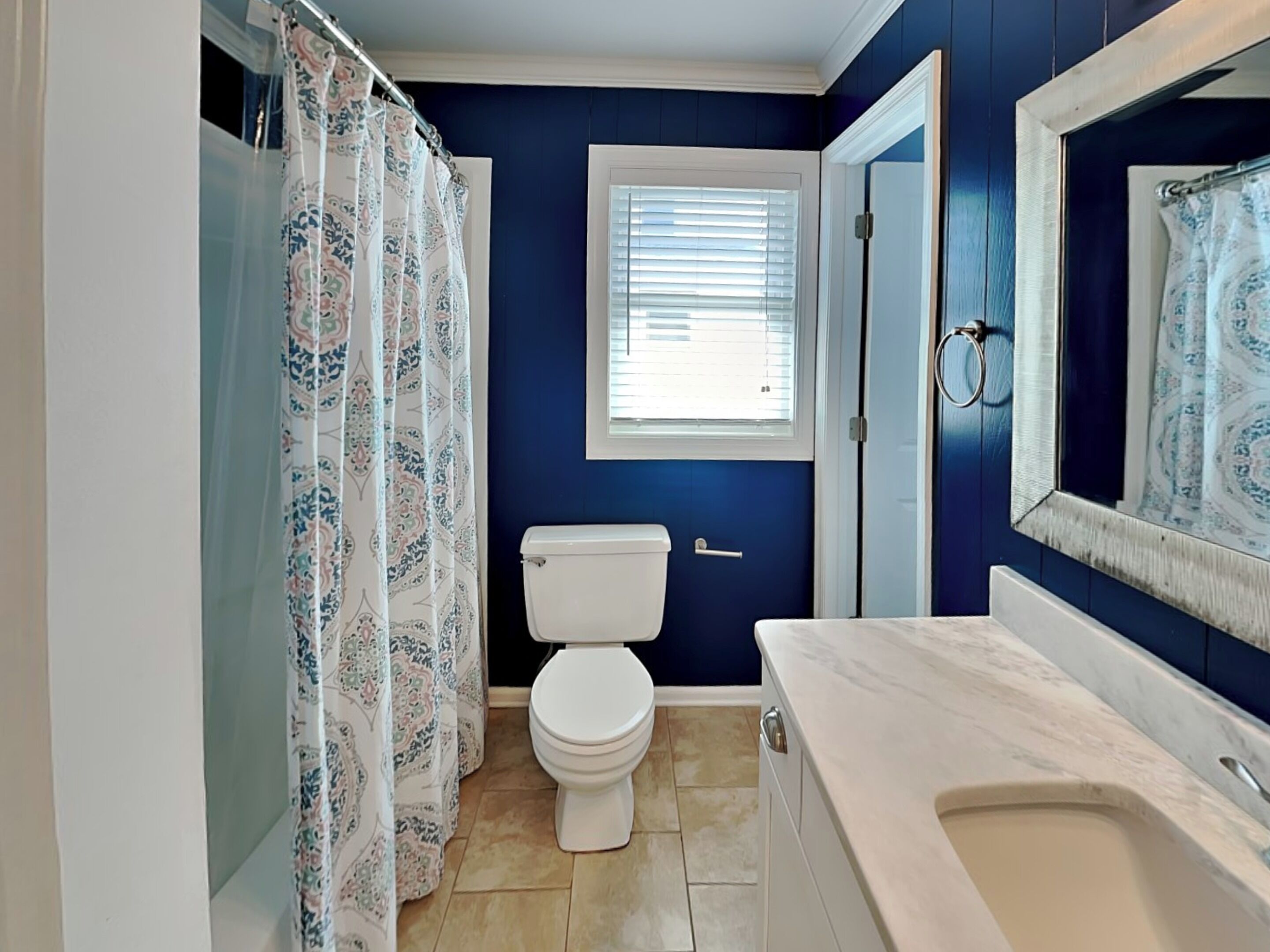 A bathroom with a toilet, a shower with a patterned curtain, a window with blinds, and a bathtub.