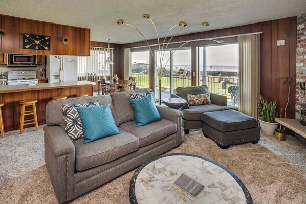 A modern living room with a sofa, ottoman, and coffee table, a kitchen area with wooden cabinets, and a large sliding glass door to the outdoors.