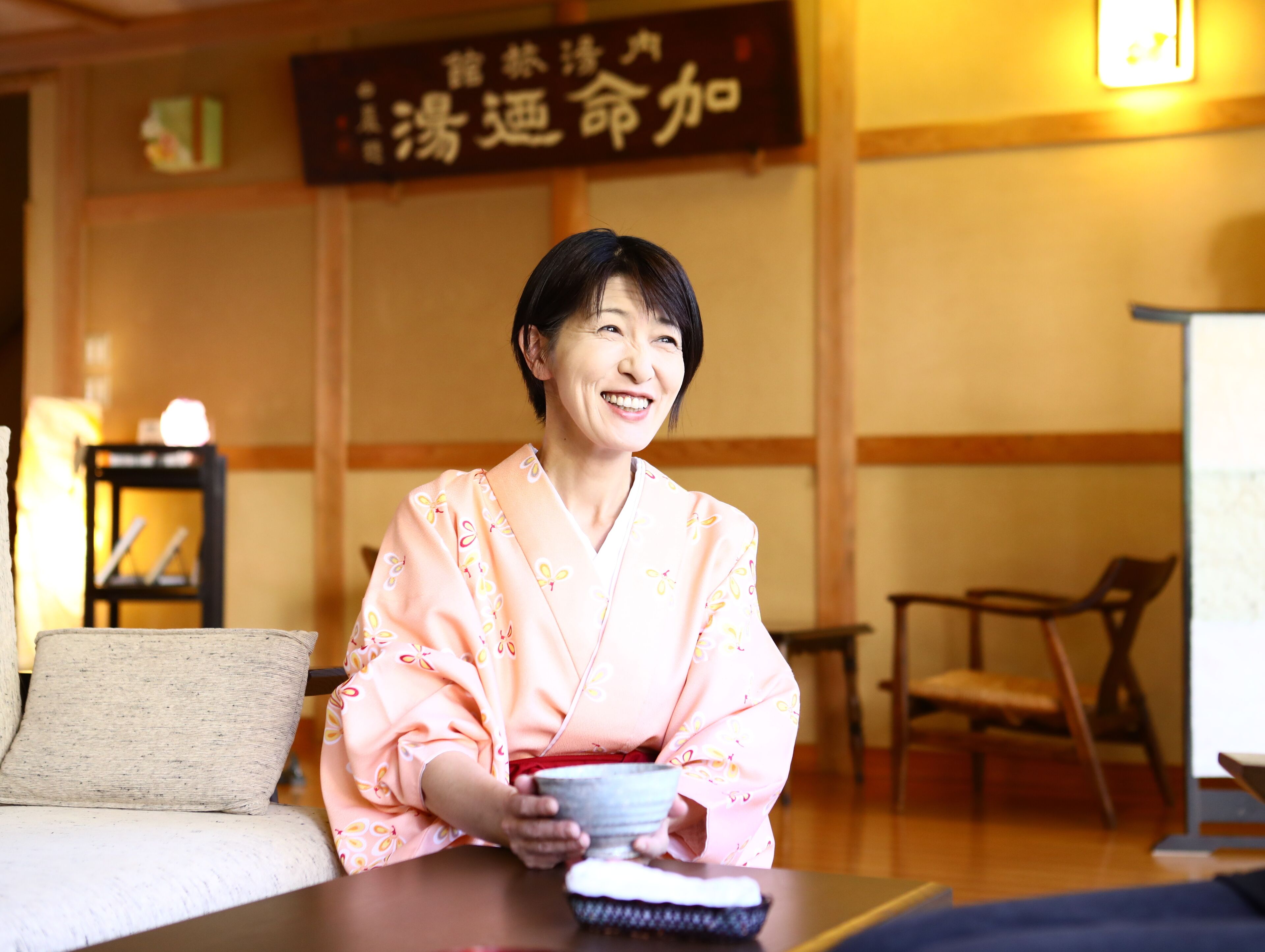 Una persona con atuendo tradicional sentada a una mesa con un tazón y un pincel, en una habitación con muebles de madera y un letrero en la pared.