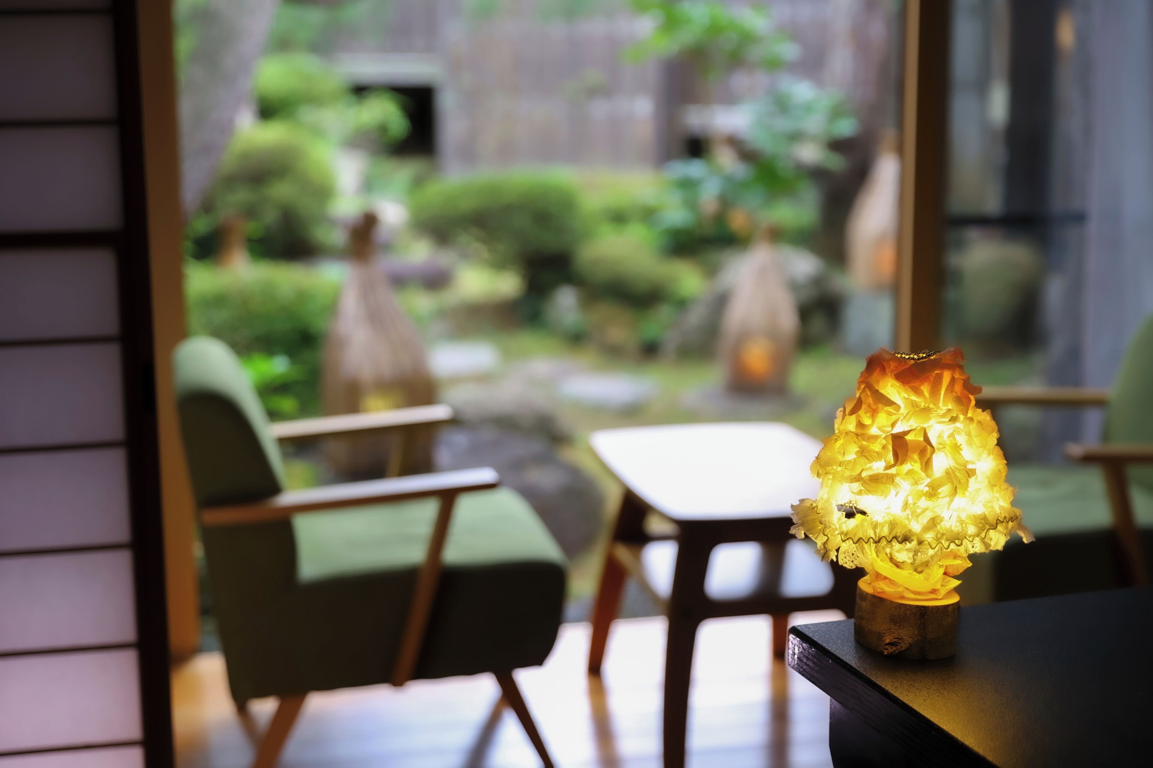 Un intérieur moderne avec une lampe de table posée sur une table en bois, des chaises vertes et une vue sur un jardin agrémenté d’une clôture en bois et de verdure.