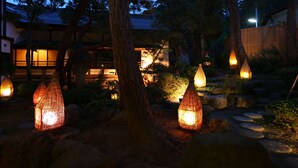 A traditional Japanese garden at dusk with illuminated lanterns, stone pathways, and a wooden building in the background. - Kamei no Yu (Yamanouchi)