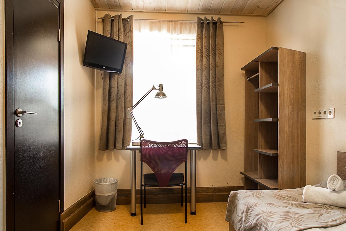 A small, furnished room with a bed, a desk with a chair, a television mounted on the wall, and a wardrobe.