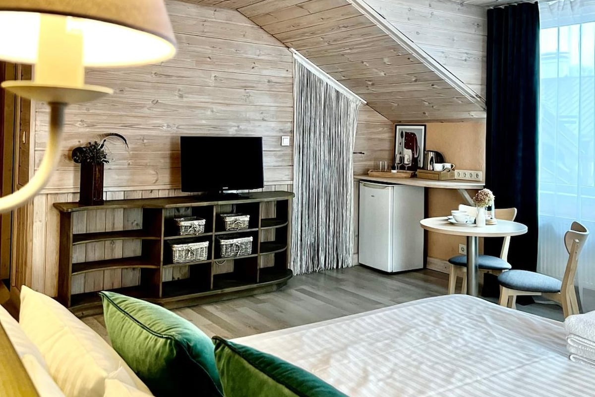 A modern bedroom with a wooden ceiling, a bed with green and white pillows, a TV, a small kitchenette, and a dining area.