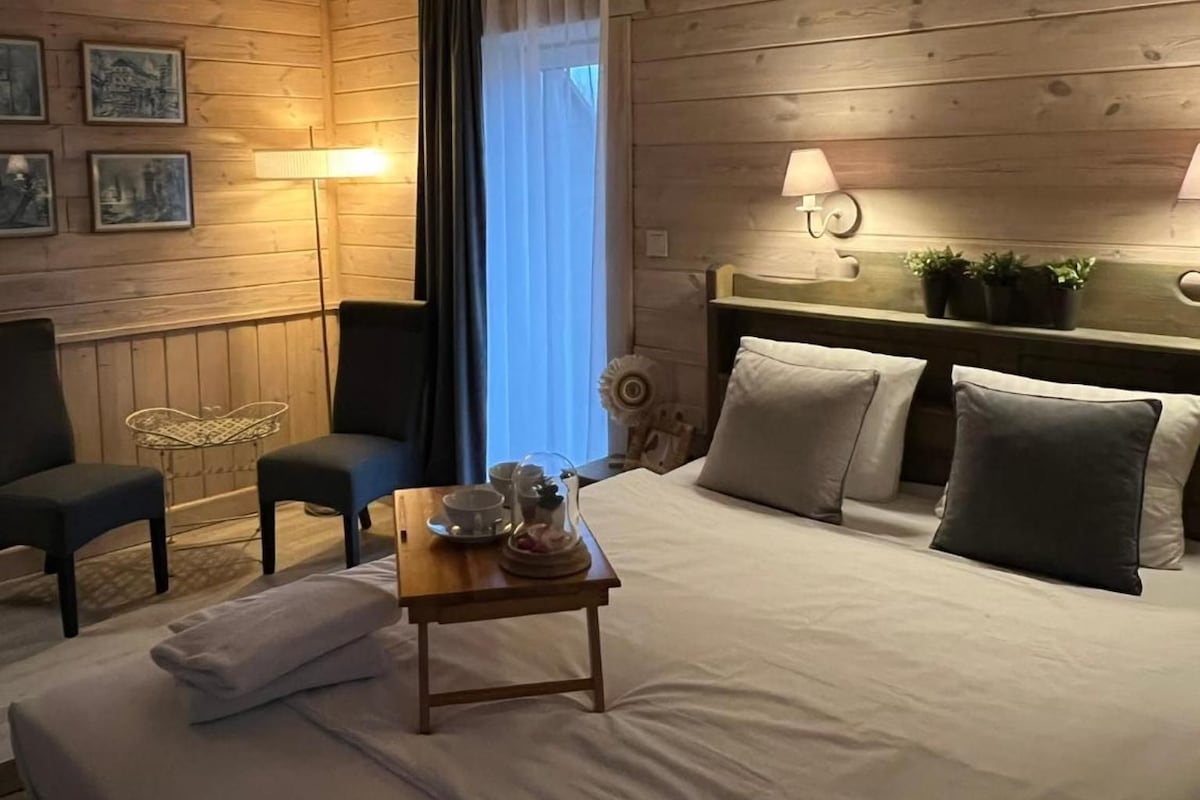 A cozy bedroom with a bed, two chairs, a small table with a cup and saucer, and a headboard with plants.