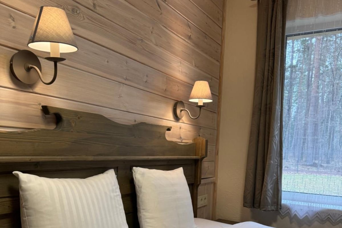 A bedroom with a wooden headboard, two wall-mounted lamps, a large bed with white bedding, and a window with curtains.