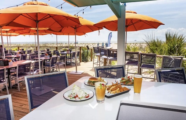 An outdoor dining area with tables and chairs under orange umbrellas, overlooking a beach.