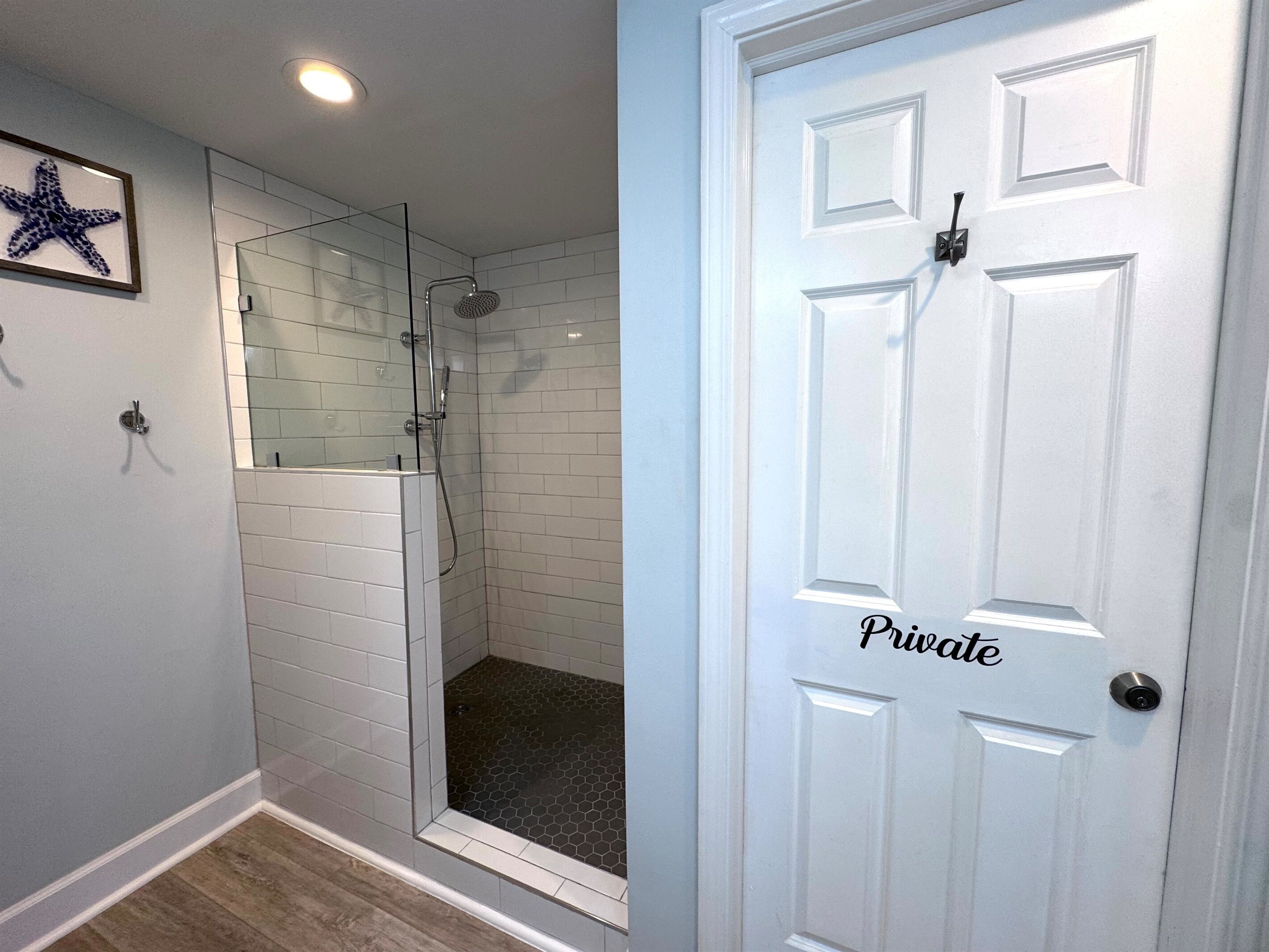 A private bathroom with a glass-enclosed shower, white tiled walls, and a wooden floor.