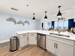 A modern kitchen with a dishwasher, double sink, and a view of the living area with a balcony. - Beach Club A1708 King/King/Queen/Sofa sleeper (Gulf Shores)
