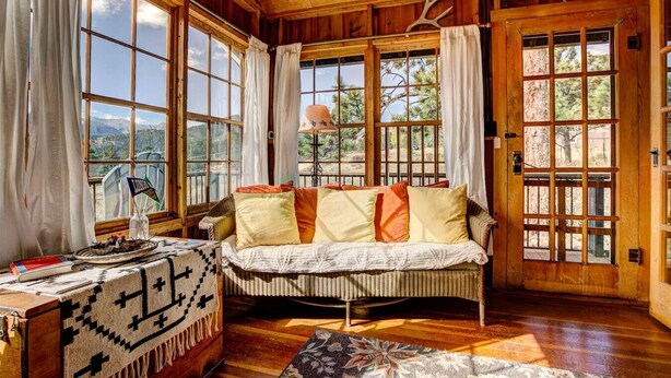 A cozy room with a wooden ceiling, large windows, a sofa with cushions, a table with a blanket, and a lamp. - Larkspur Retreat- Breathtaking Views of Longs Peak! (Estes Park)