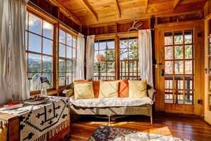 Una stanza accogliente con soffitto in legno, ampie finestre, un divano con cuscini, un tavolo con una coperta e una lampada.