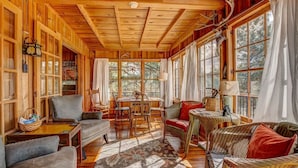 A cozy cabin interior with wooden walls, a dining area, and a large window offering a view of the outdoors. - Larkspur Retreat- Breathtaking Views of Longs Peak! (Estes Park)