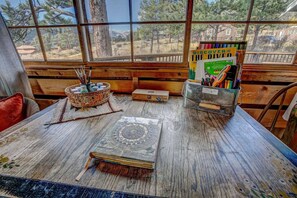 A rustic wooden table with a view of trees and a house through large windows. - Larkspur Retreat- Breathtaking Views of Longs Peak! (Estes Park)
