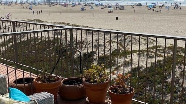 Een balkon met rieten meubilair en potplanten, met uitzicht op een strand met een pier en mensen.
