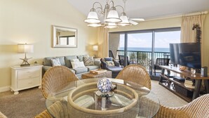 A living room with wicker furniture, a glass coffee table, a TV on a wooden stand, and a large window with a view of the ocean.