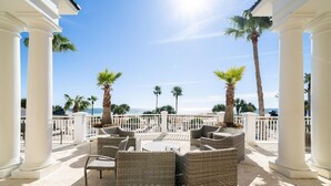 A covered outdoor seating area with wicker furniture, white columns, and palm trees.