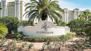 A Clubhouse sign with a palm tree in front, surrounded by greenery and a pathway.
