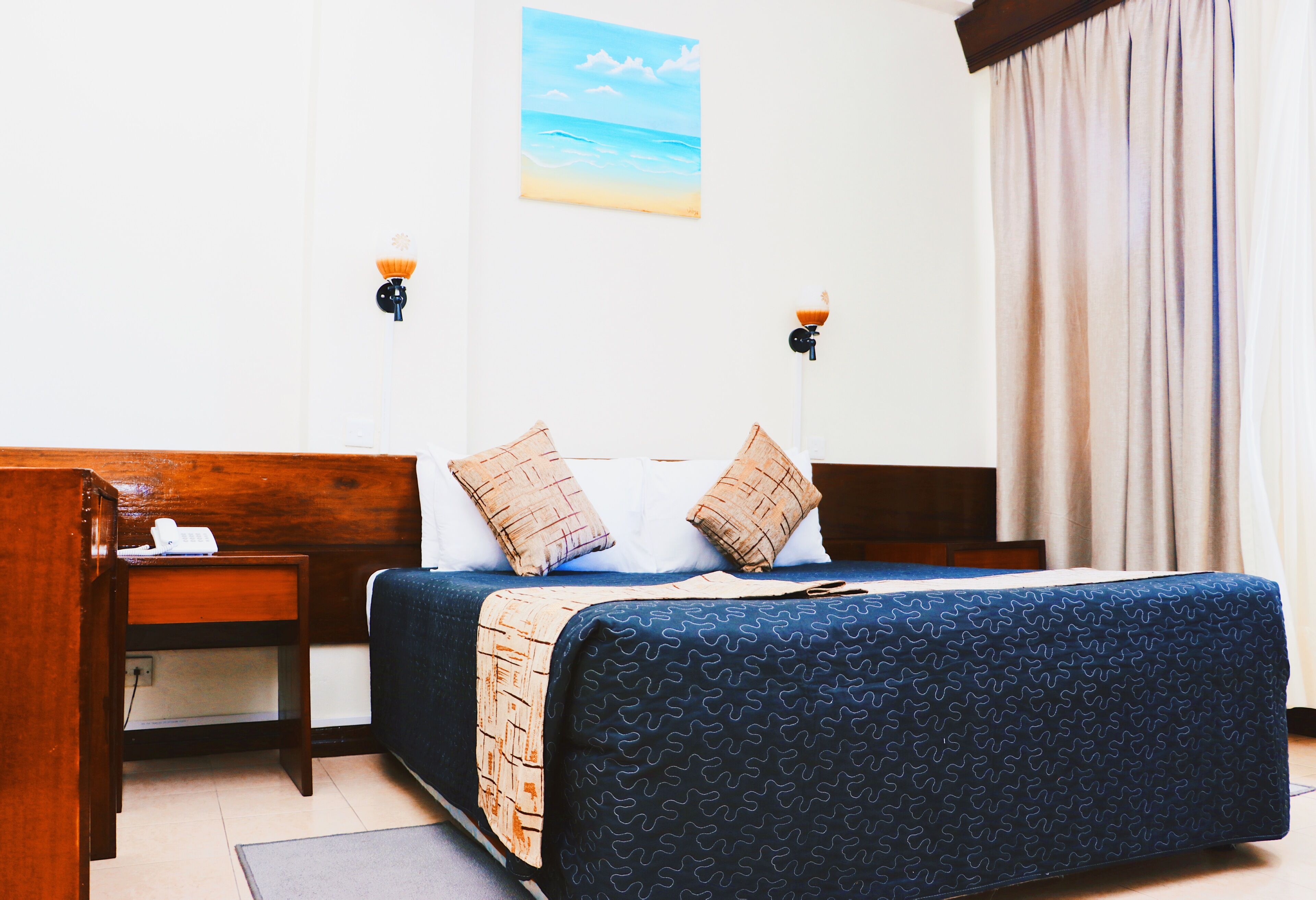 Une chambre d’hôtel avec un lit, une tête de lit en bois, des tables de chevet, une petite table, une lampe et un tableau représentant une scène de plage.