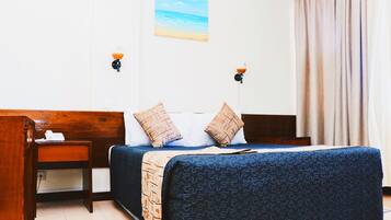 Une chambre d’hôtel avec un lit, une tête de lit en bois, des tables de chevet, une petite table, une lampe et un tableau représentant une scène de plage.