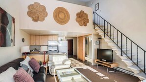 Una sala de estar con sofá, mesa de centro, televisor, escalera, cocina y decoración mural.
