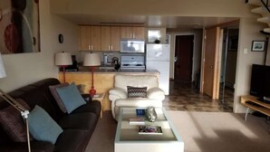 Una sala de estar con un sofá, una mesa de centro y un mueble para televisor. Se aprecia una cocina con armarios y electrodomésticos al fondo.