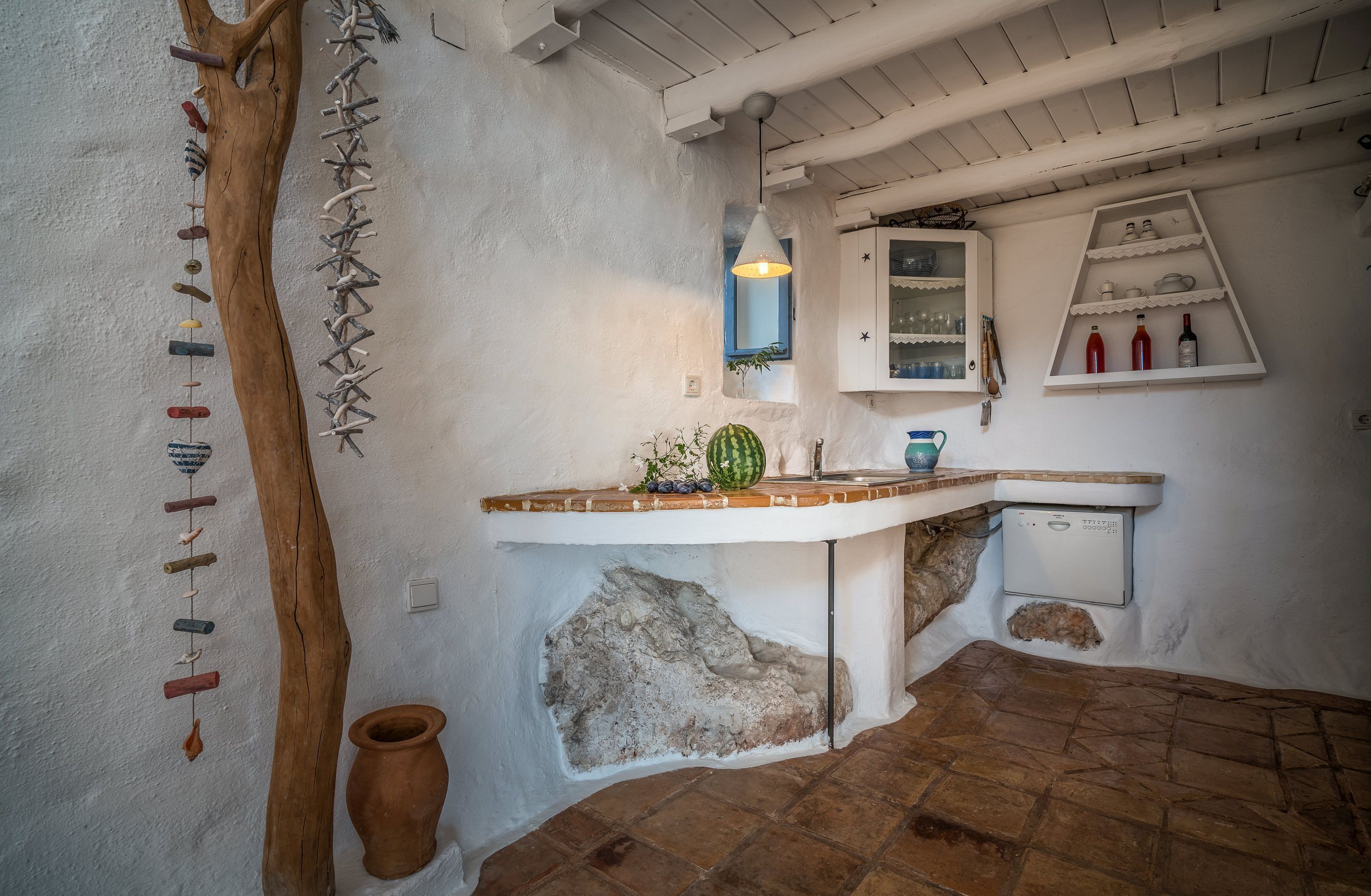 Una cocina rústica con un tronco de árbol de madera, un móvil de viento colgante, una maceta de terracota y un armario blanco con estantes.