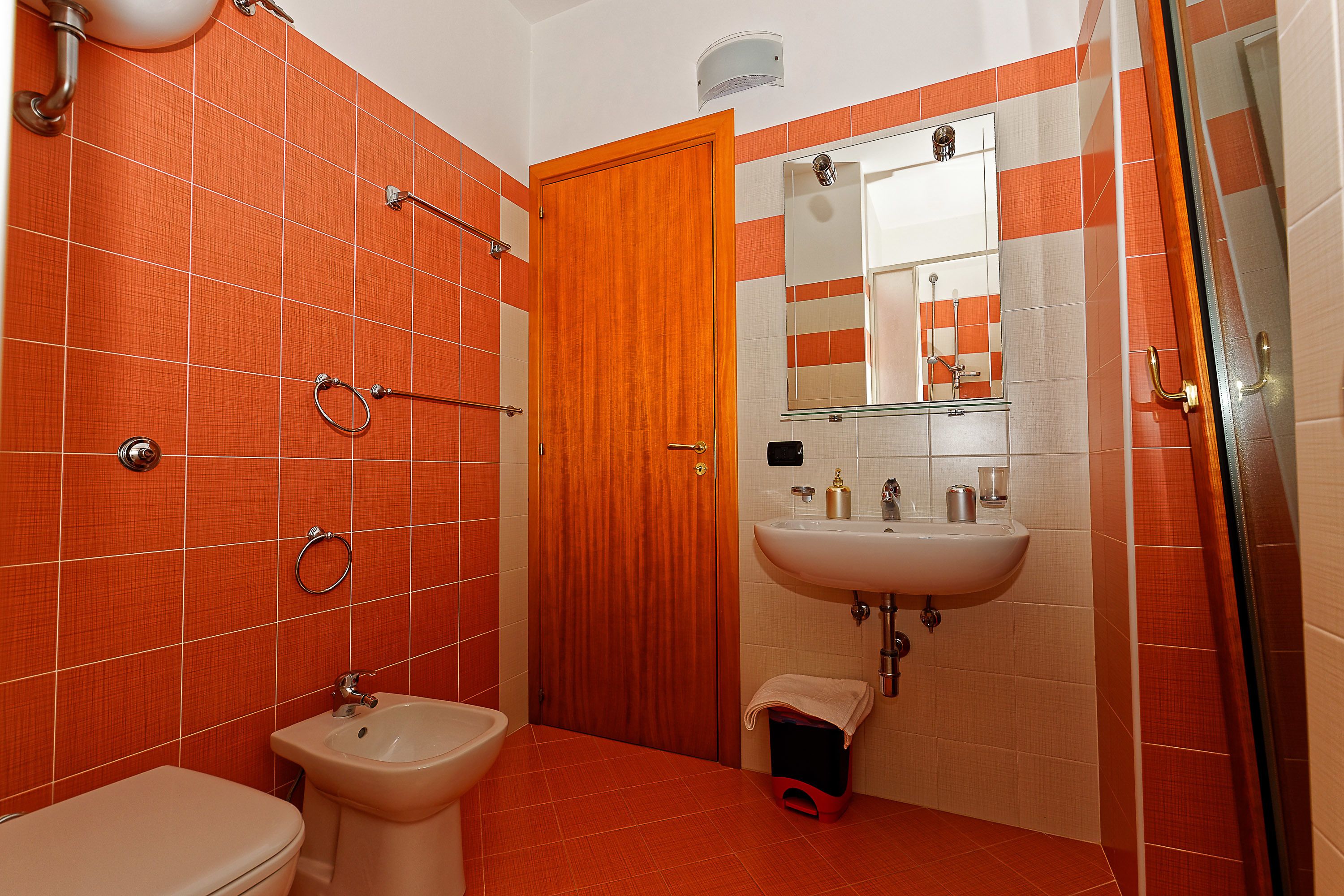 A bathroom with orange tiles, a wooden door, a sink, a toilet, and a shower area.