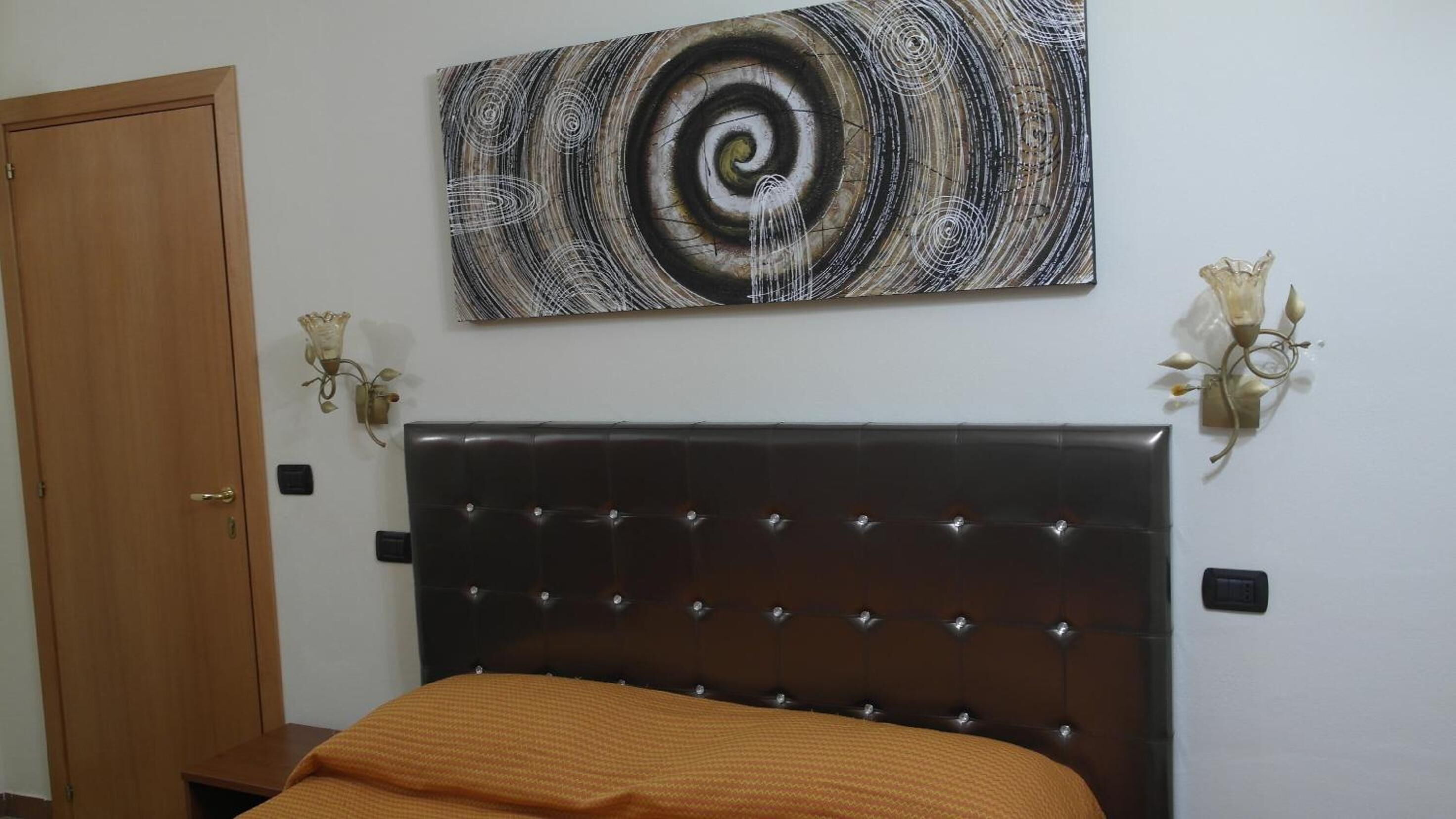 A bedroom with a dark headboard, a bed with an orange blanket, a wooden door, and a wall-mounted abstract painting.