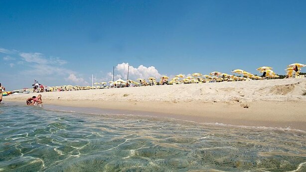 Una playa con aguas poco profundas y cristalinas, sombrillas y gente.