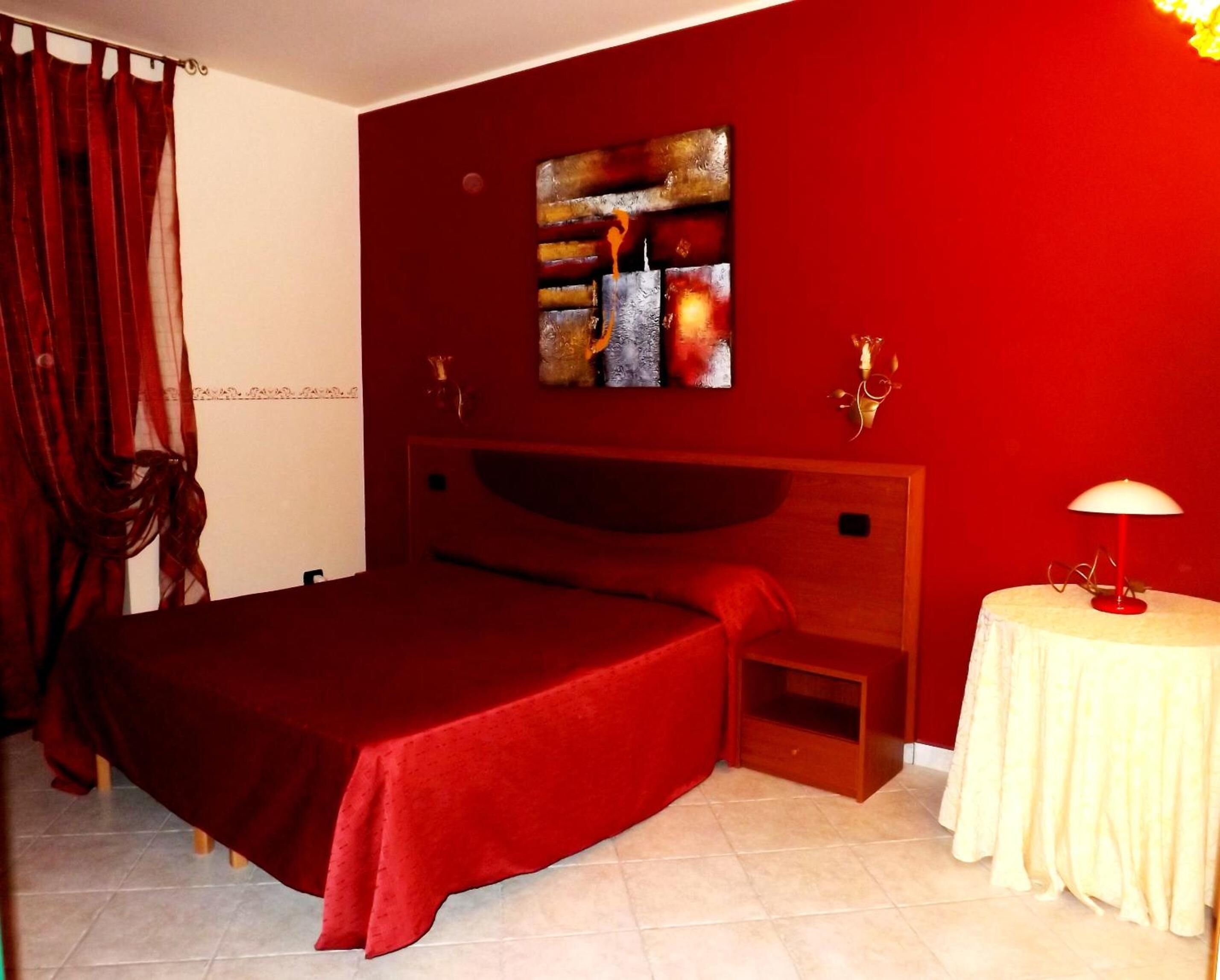 A bedroom with a red bedspread, a wooden headboard, a bedside table with a lamp, and a painting on the wall.