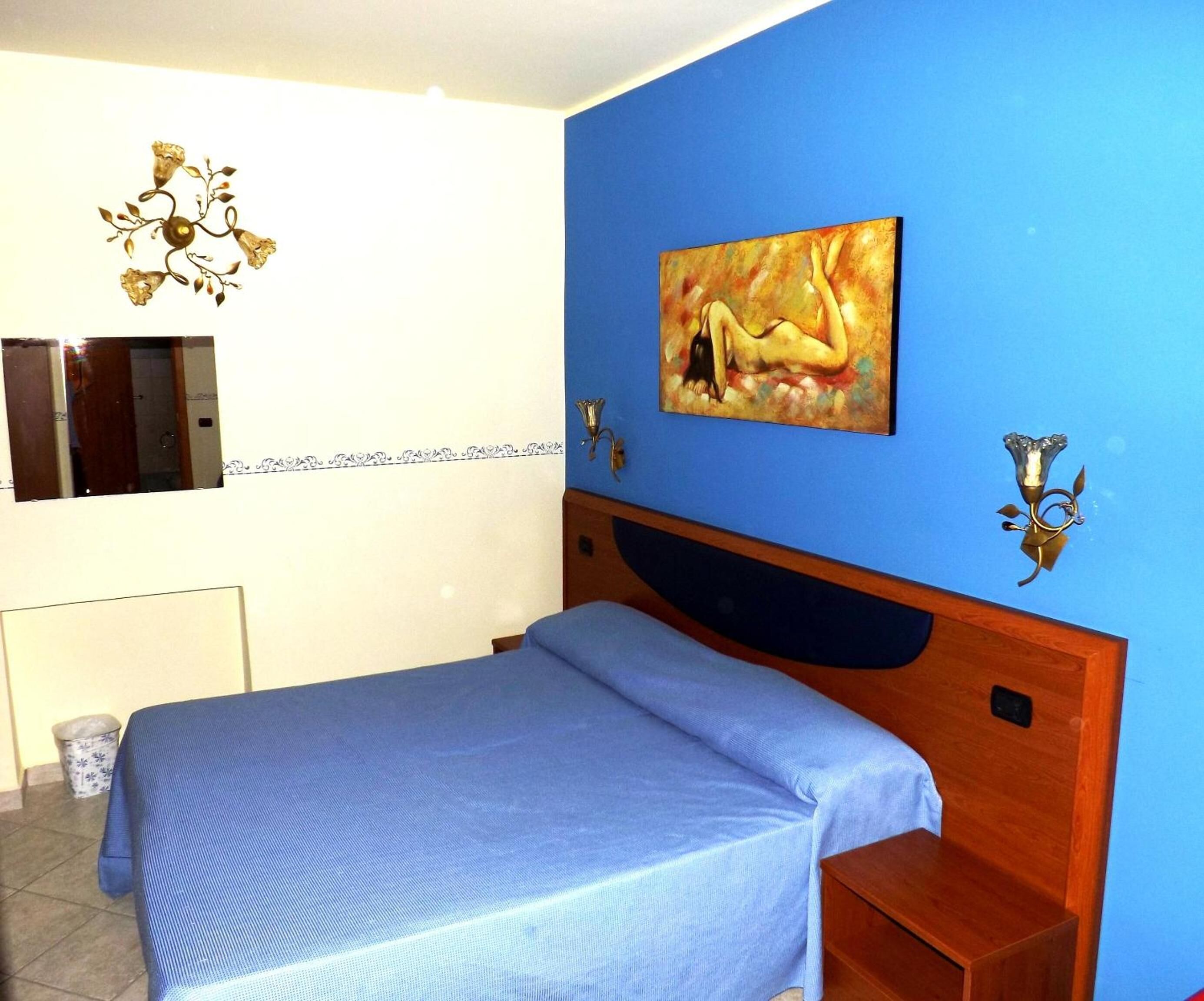A bedroom with a blue wall, a bed with a blue bedspread, a painting, a mirror, and a decorative wall sconce.