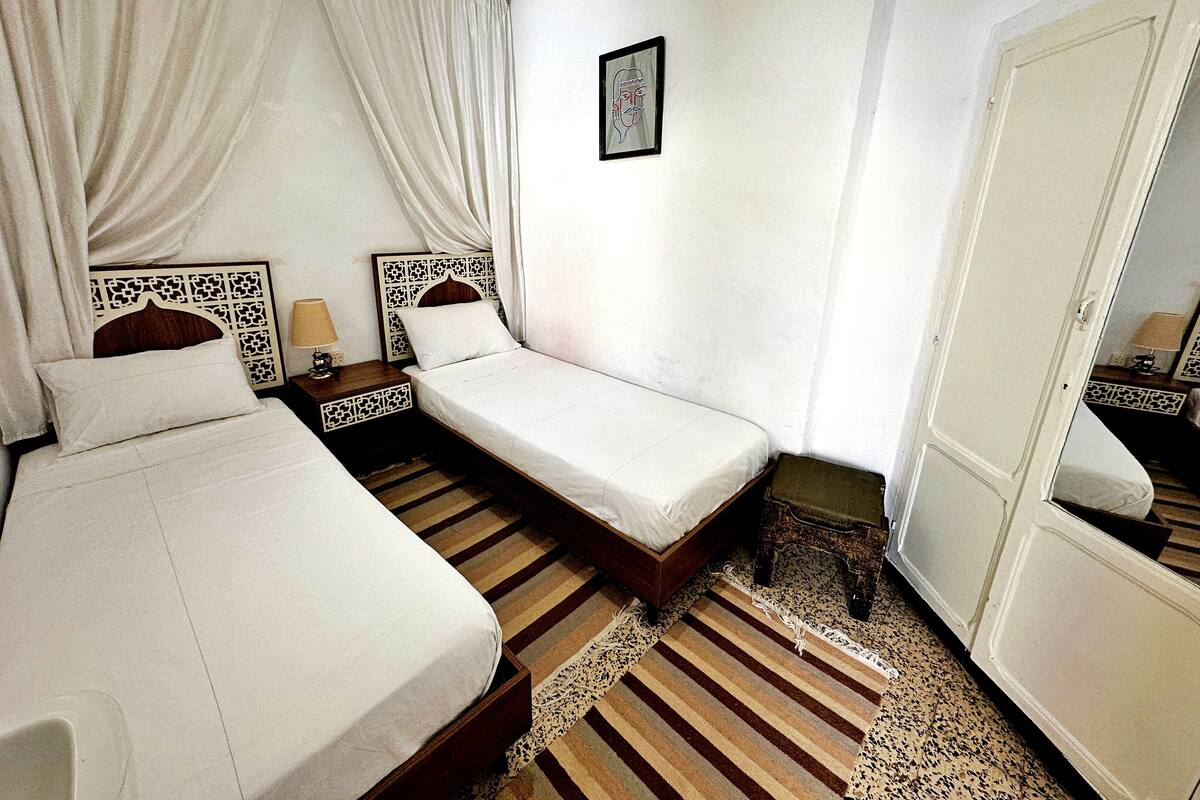 A hotel room with two single beds, a wooden floor, a mirror, and a small table with a lamp.