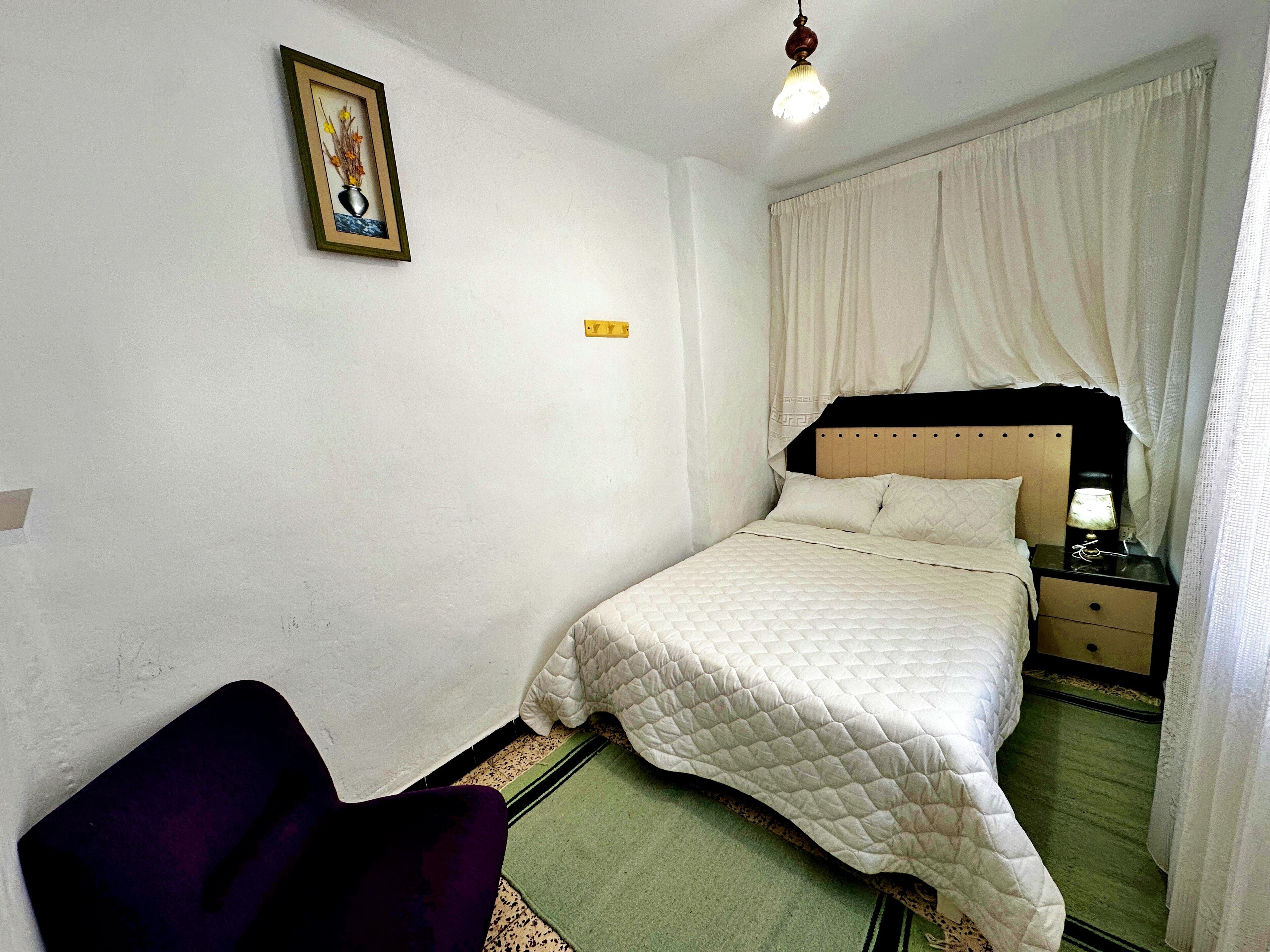 Un dormitorio con cama, mesita de noche, lámpara, silla y un cuadro en la pared.