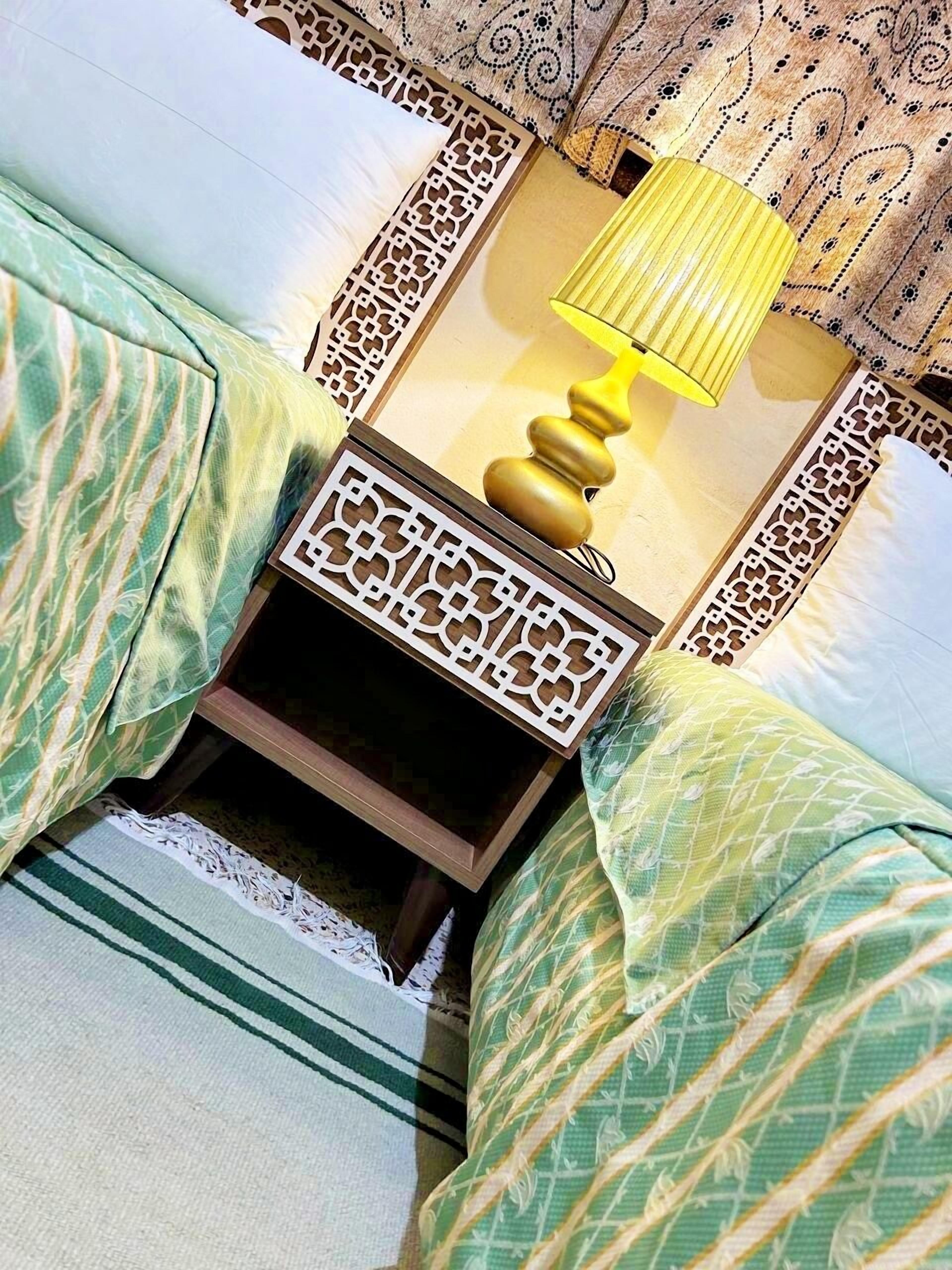 Una cama bien hecha con ropa de cama verde estampada, una mesita de noche de madera con una lámpara decorativa y un papel tapiz estampado.