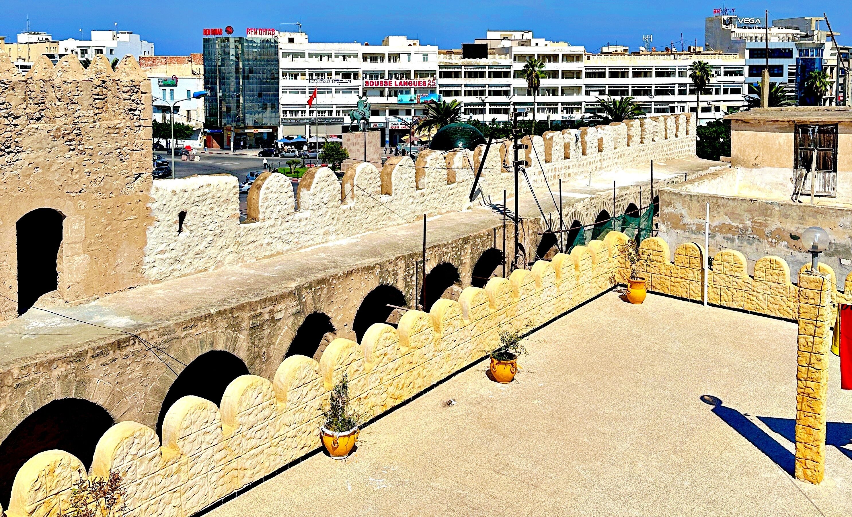 Un mur de pierre ancien, orné d’arches, une zone pavée agrémentée de plantes en pot et, au fond, des bâtiments modernes.