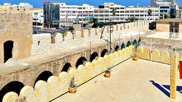 Un mur de pierre ancien, orné d’arches, une zone pavée agrémentée de plantes en pot et, au fond, des bâtiments modernes.