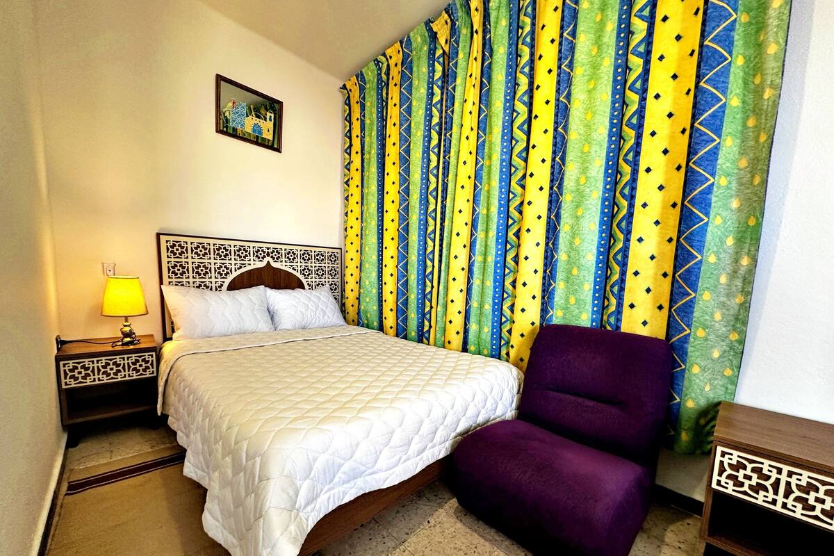 A neatly arranged hotel room with a bed, a purple armchair, a nightstand with a lamp, and a patterned curtain.