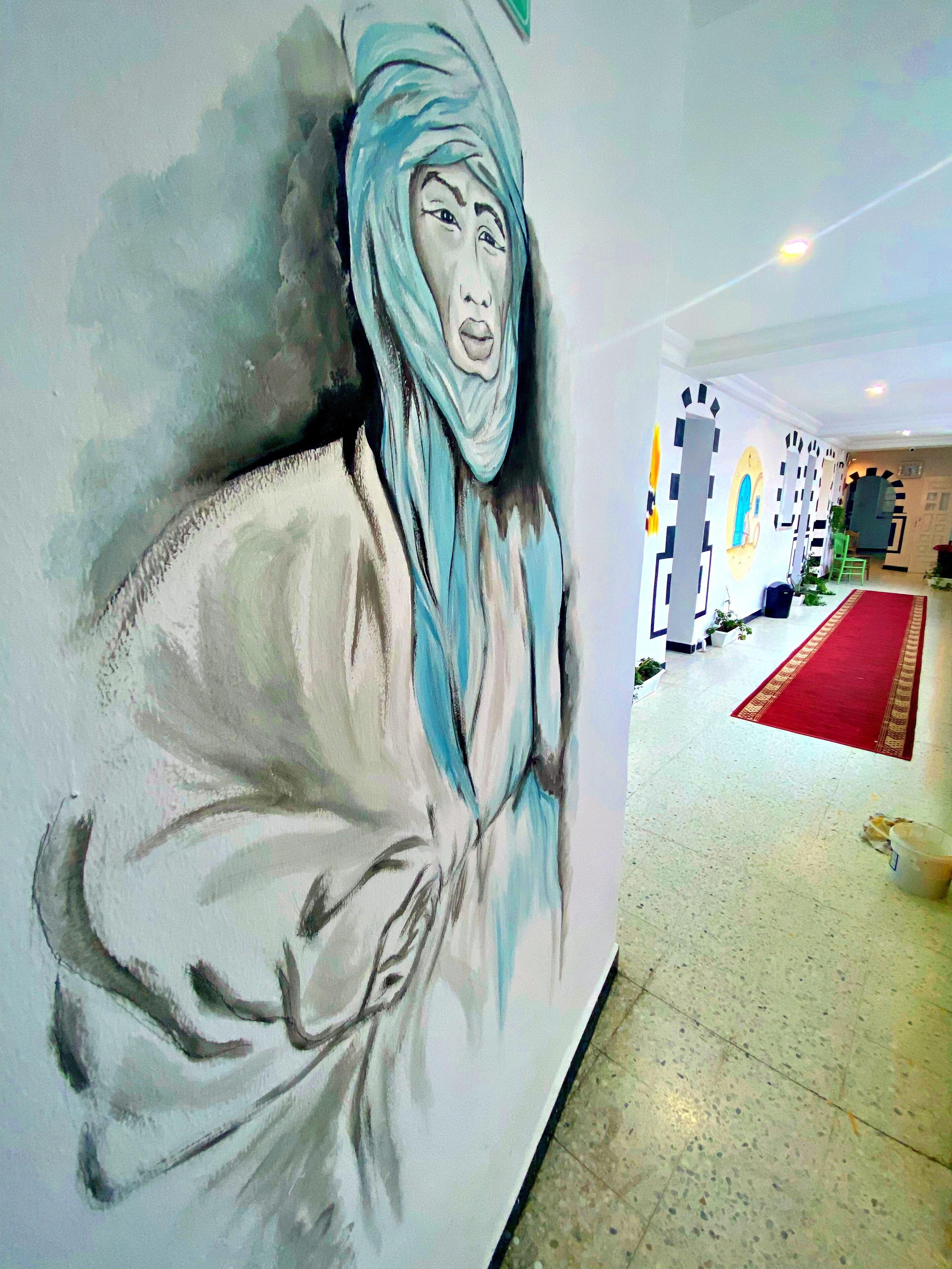 A corridor with a mural of a person in traditional attire, a red carpet, and a tiled floor.