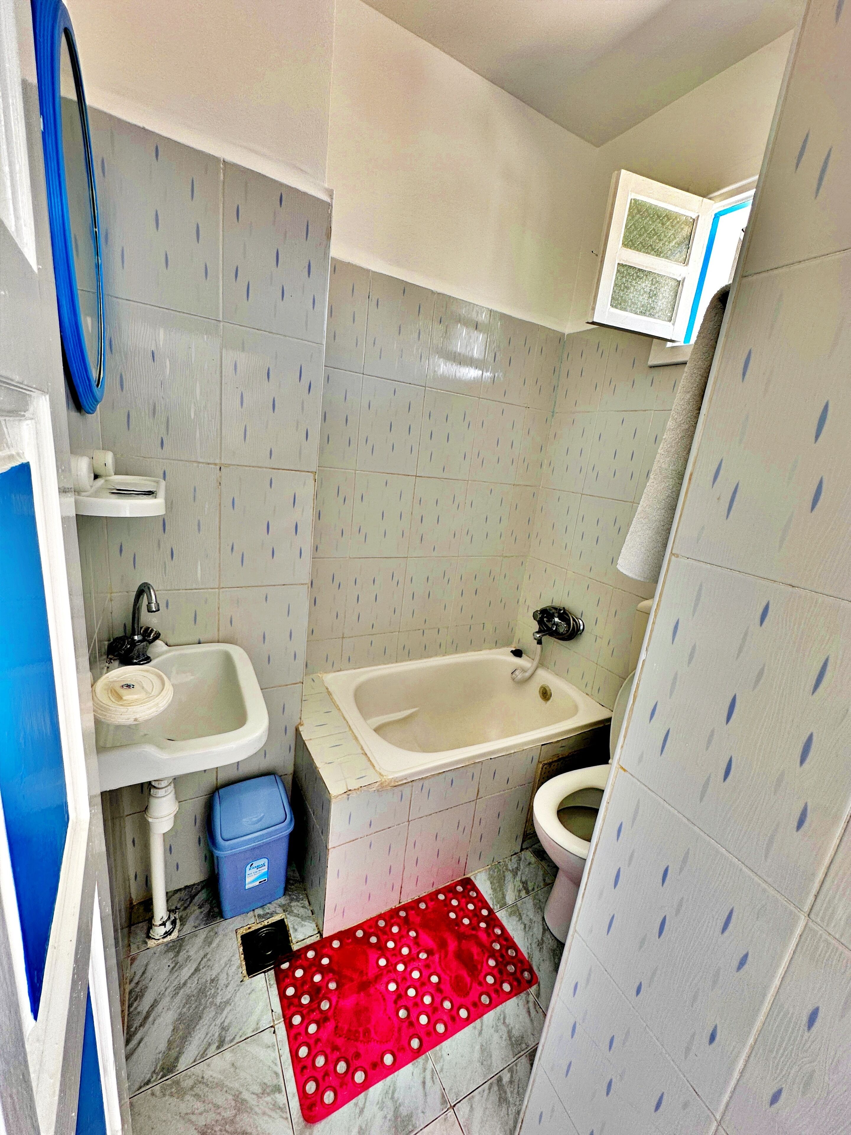 A small bathroom with a sink, toilet, and bathtub. There is a blue trash bin and a red mat on the floor.
