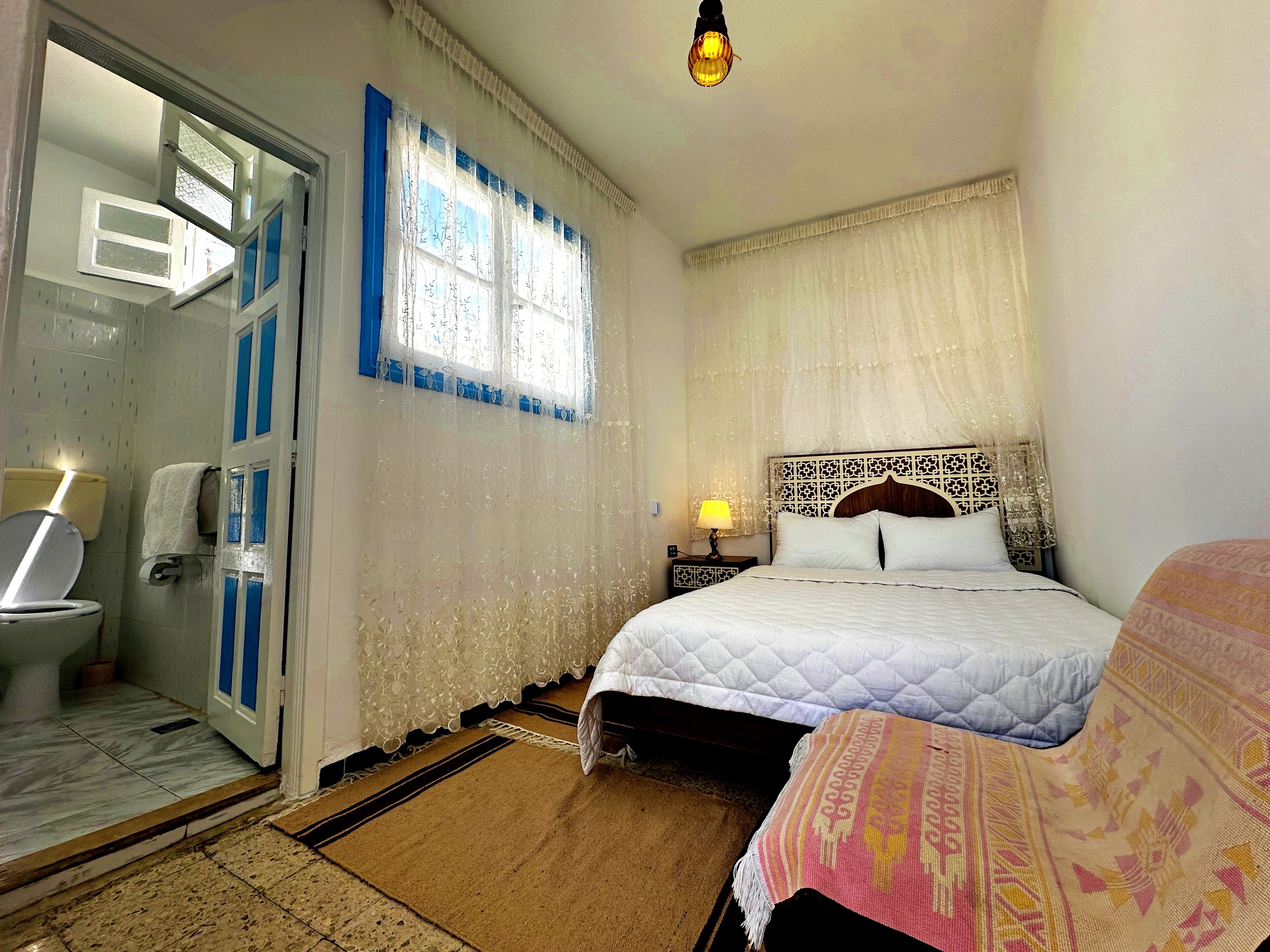 Un dormitorio con cama, mesita de noche con lámpara, ventana con cortinas y baño con inodoro y lavamanos.
