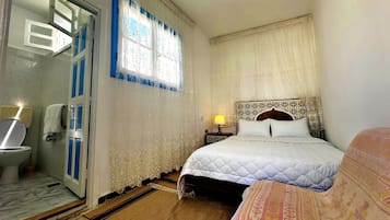 A bedroom with a bed, a bedside table with a lamp, a window with curtains, and a bathroom with a toilet and sink.