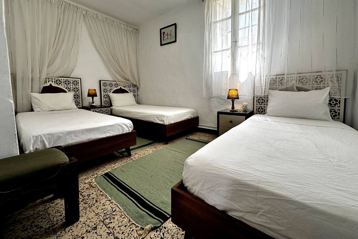 A hotel room with two single beds, a bench, a small table, and a nightstand with a lamp.