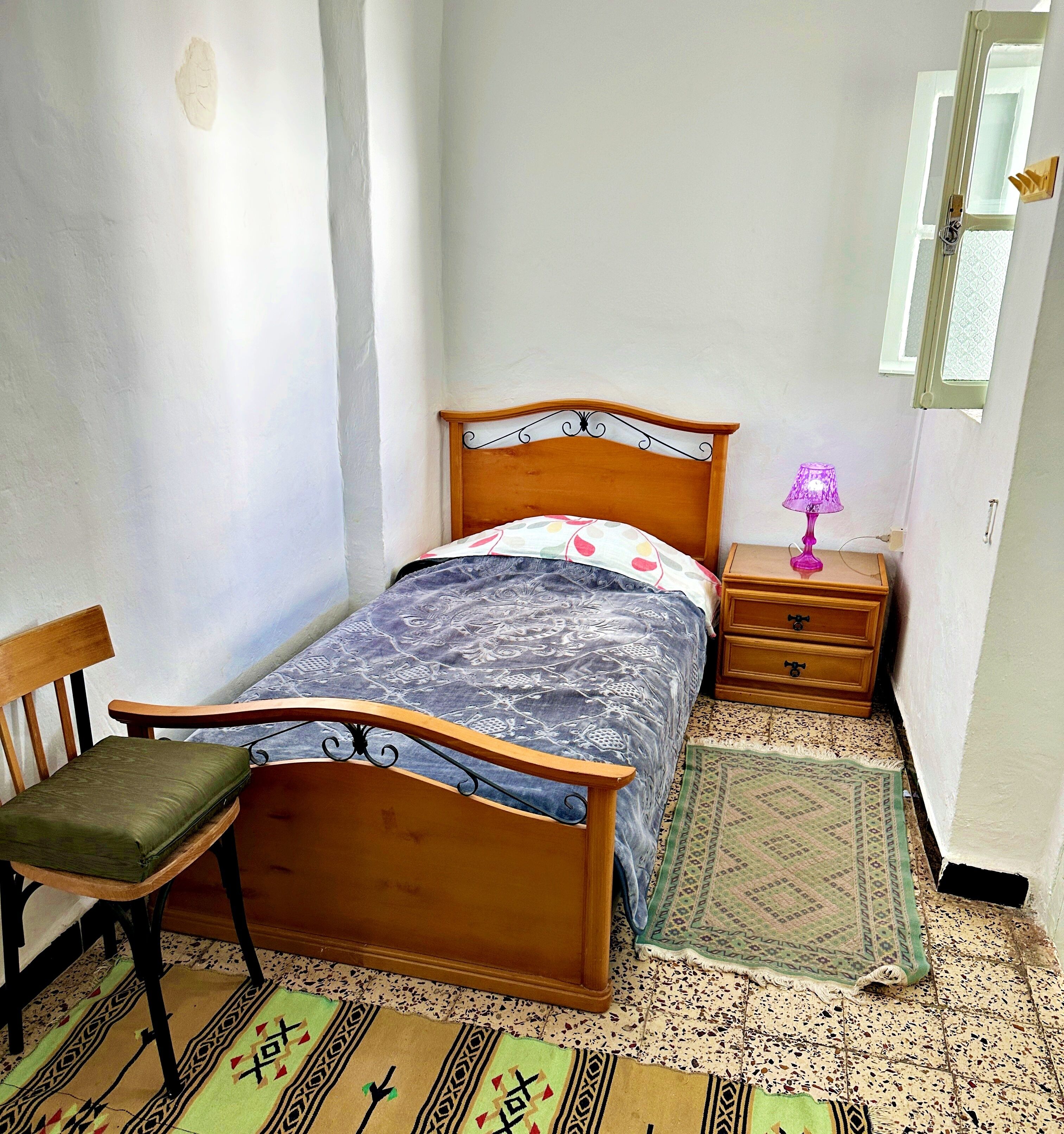 A small, neatly arranged bedroom with a single bed, a chair, a nightstand, and a lamp.