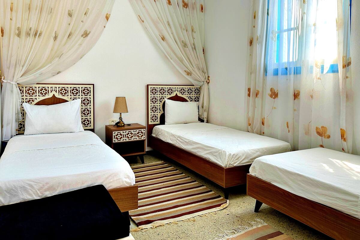 A hotel room with three single beds, a wooden headboard, a nightstand with a lamp, and a striped rug.